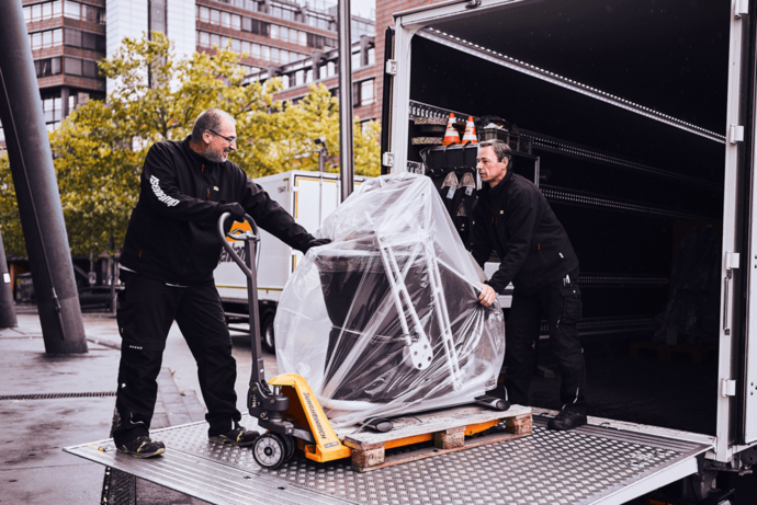 Zwei Mitarbeiter von hasenkamp schieben eine in Plastikfolie verpackte Maschine aus einem LKW-Anhänger.